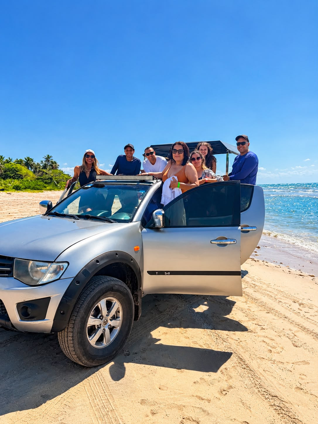 Grupo sorrindo em uma caminhonete 4x4 prata na areia da praia, com o mar ao fundo