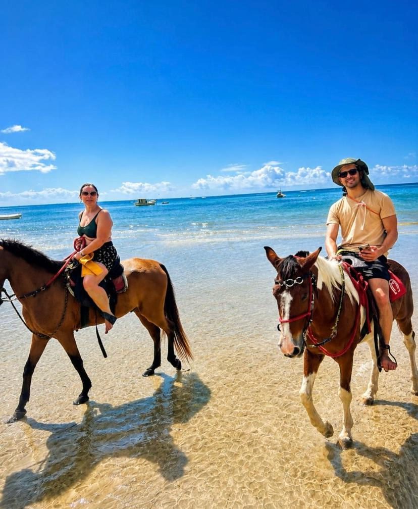 Duas pessoas sorrindo montadas a cavalo à beira-mar